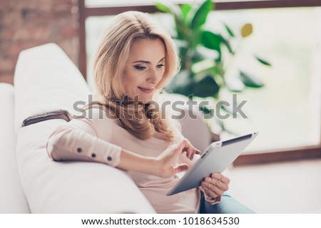 Concept of coziness and leisure at home. Close up photo of charming beautiful woman with blonde hair, she is using her digital tablet for watching photos and videos
