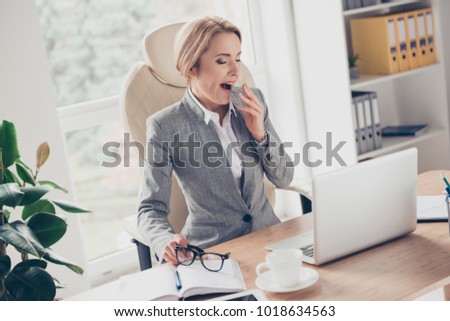 Charming, mature, middle age woman in jacket sitting at her desk in workstation, put off glasses, yawn, closing her open mouth with palm