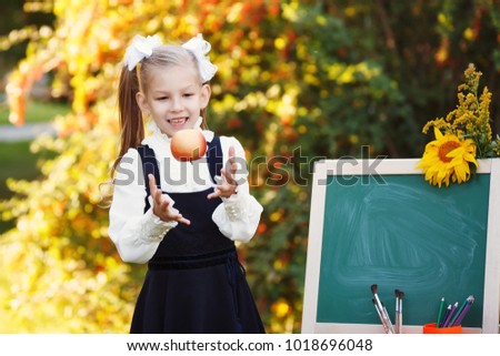 Cute schoolgirl in school uniform with a chalkboard in the autumn park. Girl looking an apple near the easel blackboard.