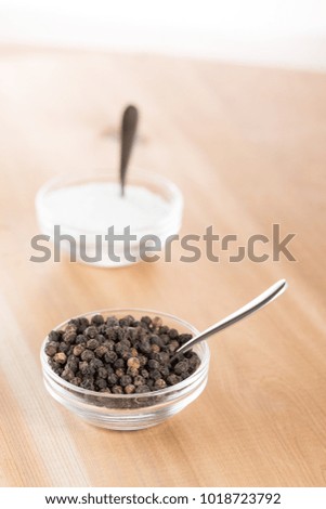 Overhead on a glass dish of round black peppercorns, with a silver teaspoon, and salt container in the blurry background, on a wood table