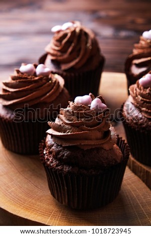 Delicious dark chocolate buttercup cupcakes, sugar heart topping, wooden textured table. Cocoa bakery, rustic wood tree slice coffee shop hipster tabletop. Obesity. Black wall background, close up.