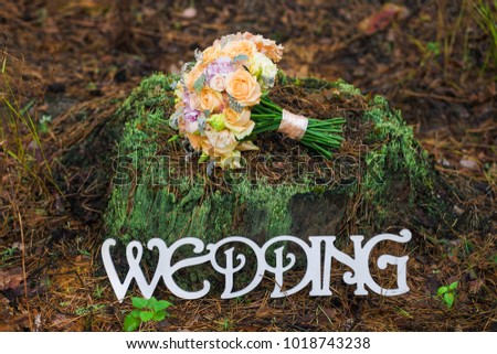 Bouquet of bridal wedding flowers on top of old stump. Decorated place in old autumn wood for wedding ceremony. Rustic style of elements of decor. White wooden word Wedding. Horizontal color photo.