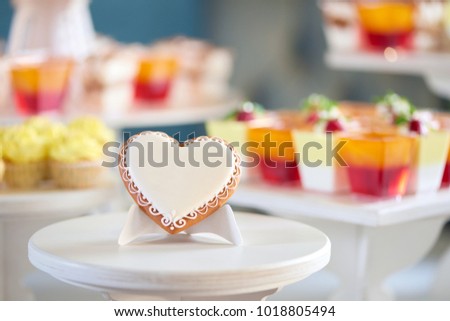 Close-up of little candy sweatheart,covered with white glaze and decorated with pattern, which stands on the wooden stand, near a yellow cupcakes and candybar on the background. Good gift for guests. Royalty-Free Stock Photo #1018805494