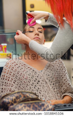 Close up of beautiful face of young woman getting make-up. The artist is applying eye mascara on her eyes. The lady closed eyes with relaxation, in a blurred background