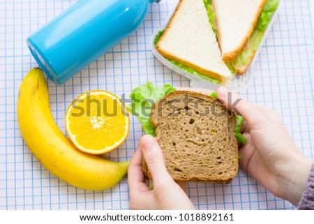 Fresh sandwich with lettuce in a plastic container, orange, a blue thermos bottle and a man's hand holding a rye bread sandwich, top view, white squared paper background