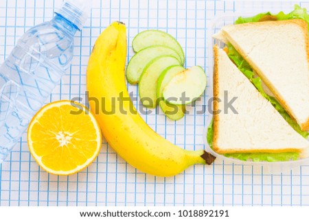 Fresh sandwich with lettuce in a plastic container, banana, orange and a bottle of still water top view, white squared paper background