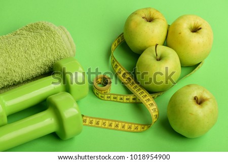 Barbells made of plastic near juicy green apples. Diet and sport regime concept. Dumbbells in bright green color, measure tape, towel and fruit on green background. Sports and healthy regime equipment Royalty-Free Stock Photo #1018954900