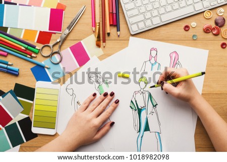Top view on fashion designer at work. Female hands drawing clothes sketch at her creative workspace and using smartphone with yellow gradient color swatches on screen, top view