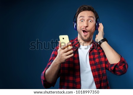 Handsome bearded man in red shirt isolated on dark blue background listening to music in his headphones on his phone and singing along with joy
