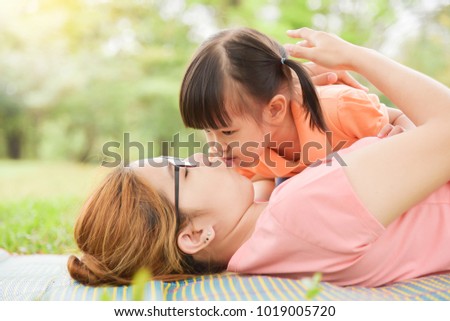 Asian little kid is kissing to her young mother while they are lying on the grass in nature at park outdoor. Family. Love.