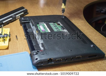 The process of dismantling the old laptop at home. A black computer rests on a cinnamon table, next to a set of screwdrivers and a battery of power. The hand unscrews the mount. Royalty-Free Stock Photo #1019009722