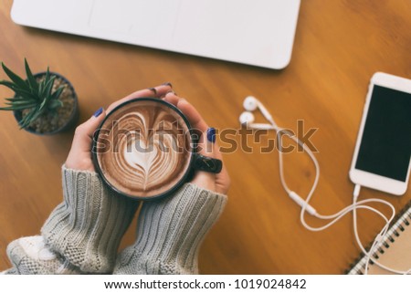 Top view female hands holding a cup of hot coffee with heart shape latte art , laptop smartphone and cactus pot on the table.