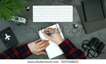 Dark table with man's hands writing in notebook and searching in web. Creative layout top view. Travel and blogger accessories: passport, camera, binoculars, notebook, keyboard. Creative concept