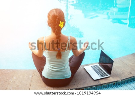 young woman freelancer meditating in lotus pose with laptop near swimming pool