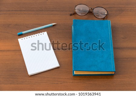 Closed book, eyeglasses, notepad and pen on wooden background