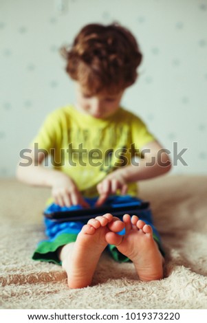 Kid with tablet sitting on bed and watching cartoons. Selective Focus on the feet. Cute boy in green t-shirt and blue pants. Vertical photo