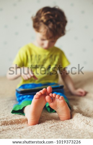 Kid with tablet sitting on bed and watching cartoons. Selective Focus on the feet. Cute boy in green t-shirt and blue pants. Vertical photo
