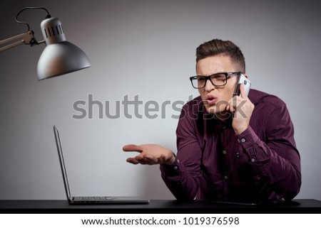  business man with glasses talking phone sits at table with noutubka on gray background, lamp, light, office, busy                              