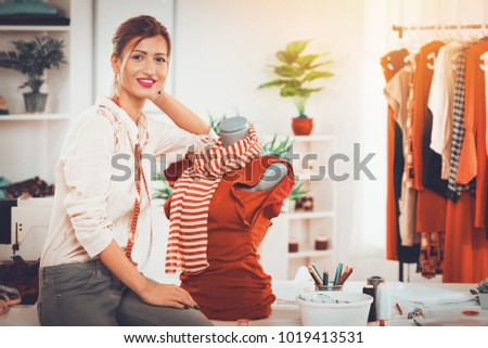 Young beautiful woman at the work place beside the dressed mannequin and with a smile looking at the camera. 