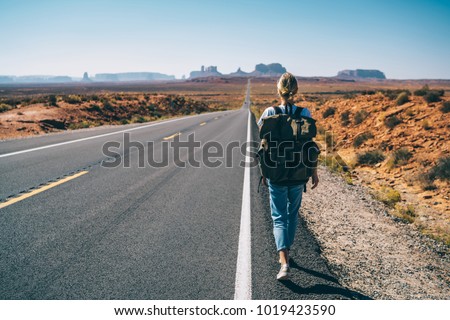 Back view of hipster girl wanderlust walking on asphalt road in wild lands on southwest getting to National park by foot, female tourist with rucksack having journey in Arizona hitchhiking on the way Royalty-Free Stock Photo #1019423590