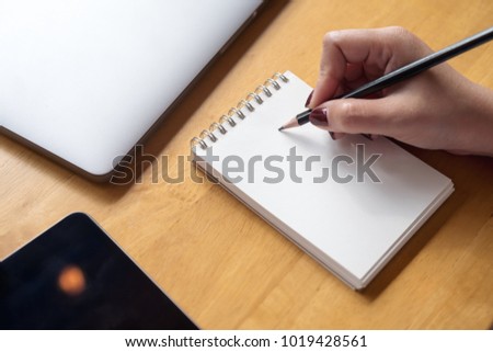 Closeup image of woman's hand writing on a blank notebook with laptop , tablet and coffee cup on wooden table background