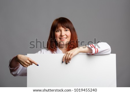 Photo of smiling chef girl in white coat pointing finger at empty sheet for inscription