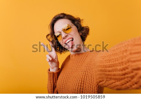 Smiling brown-haired girl posing with tongue out. Close-up portrait of adorable pretty lady in good mood making selfie on bright background.