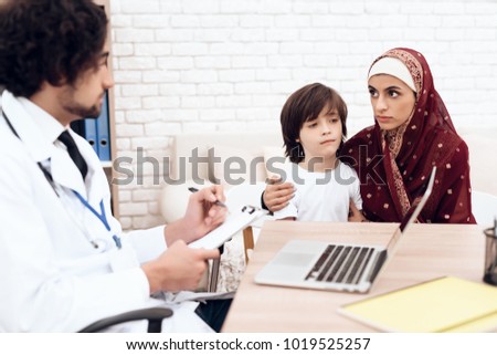 A woman hijab talks to a doctor. Near her sits a sick son, with whom she came to the hospital. The doctor on the desk is a laptop.