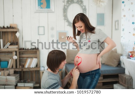 Pregnant woman with big belly. man paints heart with paints on tummy. Couple with kid clothes. Wife, husband. Pregnancy, parenthood, family, motherhood, parents, children, people, expectation concept