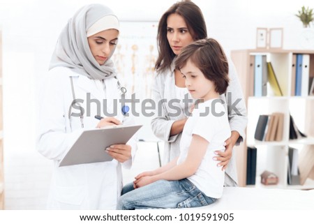 An Arab woman doctor writes a diagnosis of a sick boy. The boy came with his mother to the hospital. He has a cold.