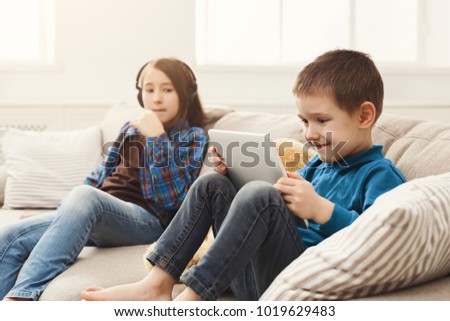 Two kids with gadgets. Sister listening to music, brother watching cartoon on digital tablet on sofa at home. Family friendship and communication concept