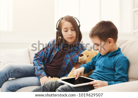 Two kids with gadgets. Sister listening to music, brother sharing funny content on digital tablet on sofa at home. Family friendship and communication concept