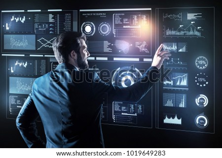 Gentle touch. Calm experienced clever programmer looking elegant in his suit and feeling happy while slightly touching a transparent screen of his futuristic device