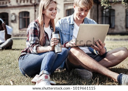 Hot tea. Delighted nice positive woman holing a thermo cup and looking at the laptop screen while sitting together with her friend on the grass