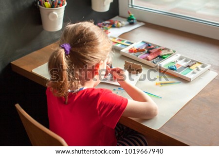 Little girl in a red and blue dress sculpts from plasticine, children's room with a slate wall, pencils and plasticine, children's creativity, a chair and a table for classes, kids craft, cartoon.