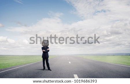 Businessman in suit with old TV instead of head keeping arms crossed while standing on the road with beautiful landscape on background.