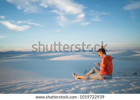 Young woman using smartphone sending pictures from trip satisfied with data connection in White Sands desert,hipster girl sitting on dune enjoying journey to New Mexico using mobile phone for chatting