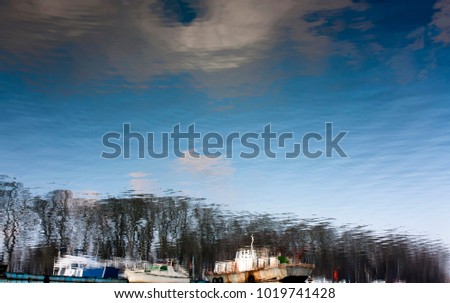 Blurry reflection of anchored old boats, trees, blue sky and clouds in river water