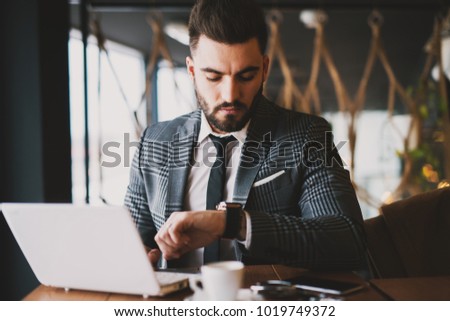 Attractive suited man checking the time on the watch while using laptop and drinking coffee in the coffee shop.  Royalty-Free Stock Photo #1019749372