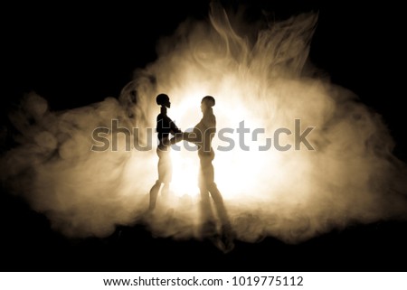 Love Valentine`s Day concept.Sillhouette of sweet young couple in love standing in the field and hugging on dark toned foggy background. Decoration with doll figures on table shot. Selective focus