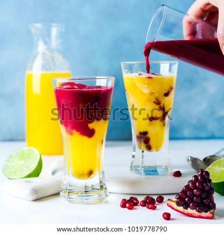 pouring grenadine into two glasses of alcoholic cocktail with orange liqueur, freshly squeezed pineapple and pomegranate juice and lime and ice on the table.