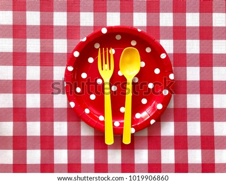 Closeup A red polka dot paper plate with yellow plastic fork & spoon on red & white checkered pattern background.The concept of party accessories,picnic utensil,Food display.Selective focus.Copy space