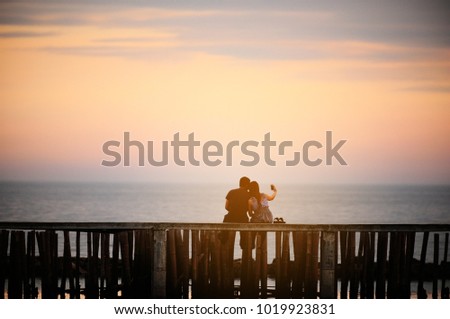 Young couple sitting on the bridge nearby the sea at sunset with flare light,Thailand. Concept for travel.
