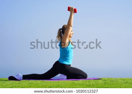 Young woman on grass with dumbbells in hands on the background of the sea. Fitness girl doing exercises with weights on the beach under the morning sun. The concept of health and weight loss.