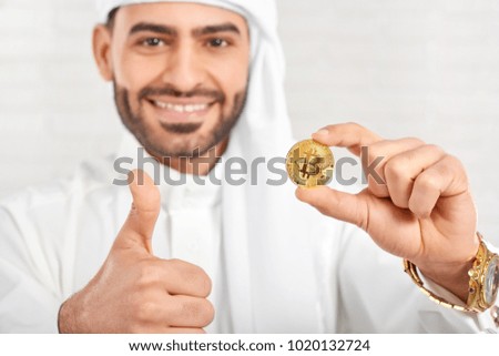 Studio close-up of a smiling Arabian businessman keeps bitcoin and looks very happy, standing on white background. He wears traditional white Arabian costume,golden watch and cufflinks.