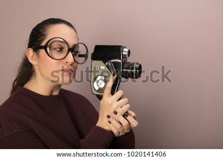 Attractive woman photographer wearing nerdy glasses peering into the viewfinder of a vintage video camera pointed towards blank copy space on a brown background