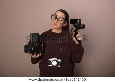 Nerdy female photography buff wearing over sized glasses with three vintage cameras holding up a video camera and staring at it with copy space