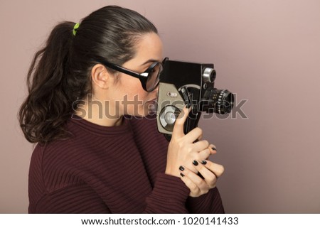 Attractive woman photographer wearing nerdy glasses peering into the viewfinder of a vintage 8mm film camera pointed towards blank copy space on a brown background