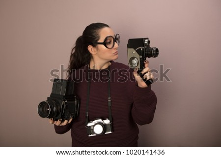 Nerdy female photography buff wearing over sized glasses with three vintage cameras holding up a video camera and staring at it with copy space