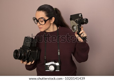 Nerdy young attractive female camera boffin with over size glasses in a stereotypical representation holding vintage stills, video and compact cameras